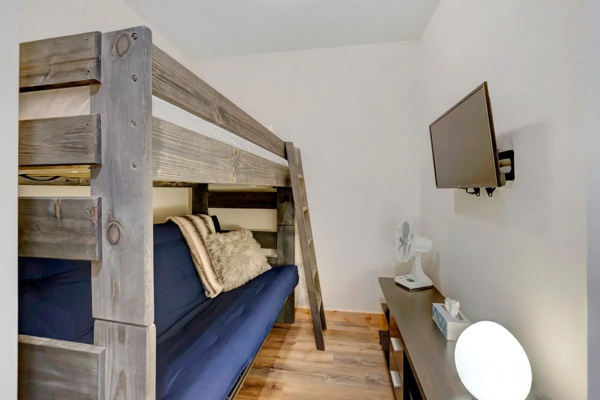 A comfortable bunk room featuring a bunk bed (twin over full), a sofa bed, television, a table lamp, and a fan. The room offers flexible, family-friendly sleeping and lounging space that caters to kids, teens, or extra guests.