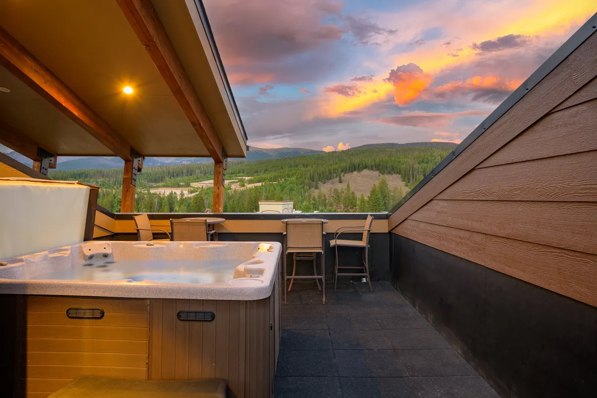 A private rooftop deck with a steaming hot tub, surrounded by comfortable outdoor seating and set against a breathtaking mountain backdrop. The sky glows with sunset hues, creating an inviting ambiance for relaxation and socializing. Patio tables and lounge chairs offer plenty of space for guests to gather and unwind.