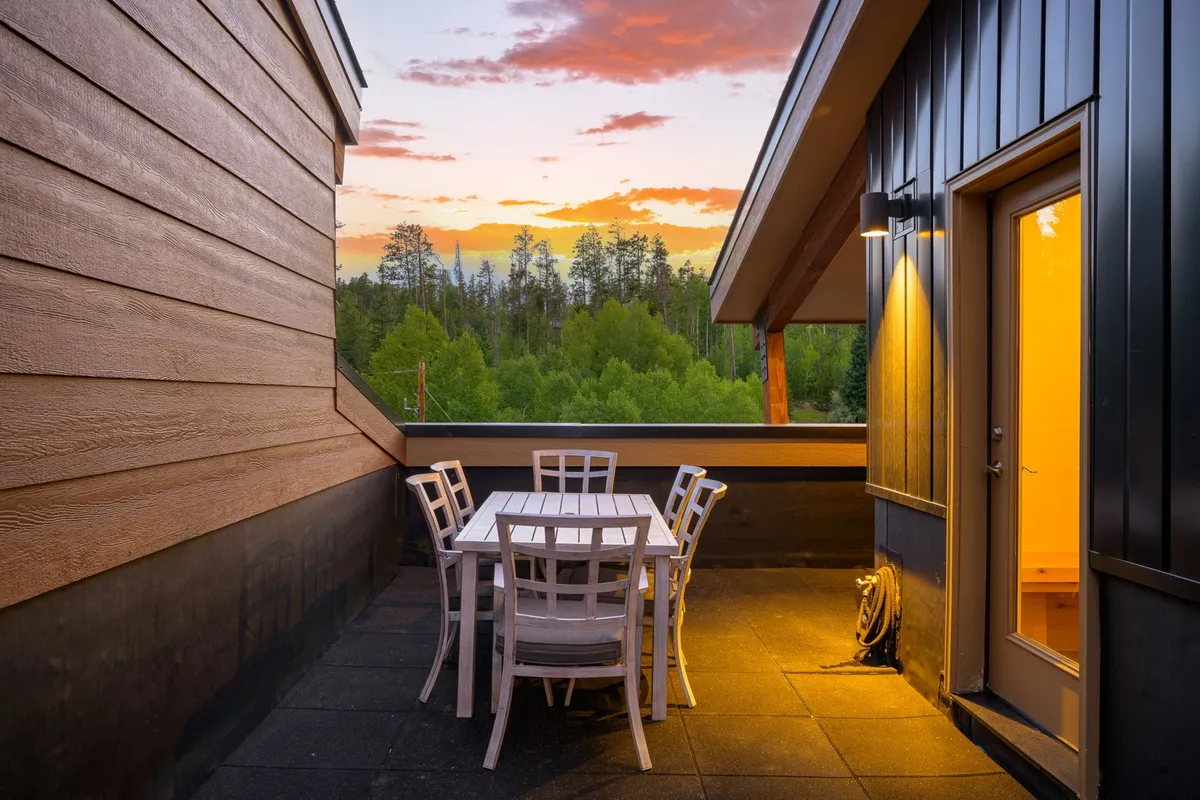 A tranquil deck scene with a spacious outdoor dining table surrounded by trees and set beneath a colorful sunset sky. The table is prepared for an evening gathering, inviting guests to share meals and stories while immersed in nature.