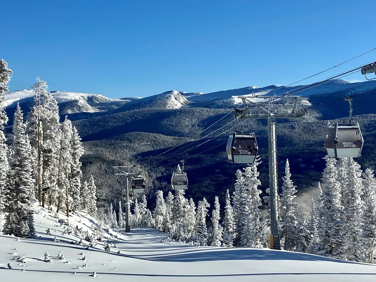 This image showcases a ski lift gliding over snow-covered trees, with a dramatic mountain range in the background. The landscape is blanketed in pristine snow, evoking the excitement and adventure of winter sports. The overall scene is vibrant and inviting, capturing the essence of winter in Winter Park.