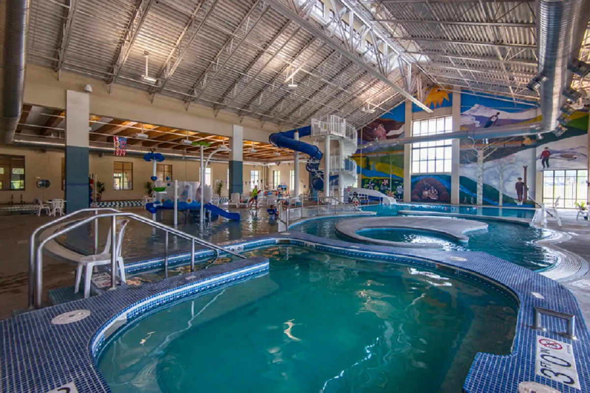 A lively indoor swimming pool featuring a colorful mural, a water slide, and children playing and swimming, surrounded by poolside seating.