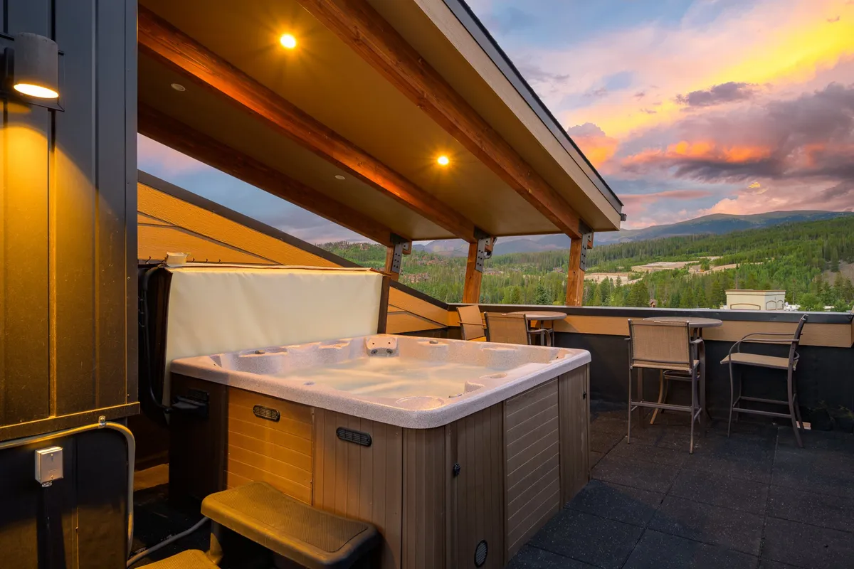 This image showcases an inviting outdoor hot tub with sweeping mountain views and an outdoor bar seating area. Friends are seen relaxing in the hot tub, enjoying drinks as the sun sets, capturing a tranquil yet social atmosphere.
