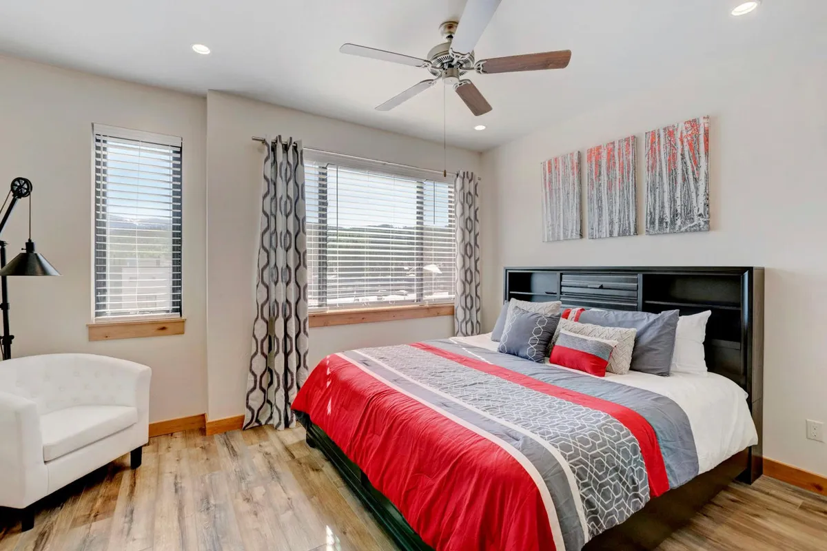 A cozy bedroom with a bed dressed in red and gray bedding, large windows allowing abundant natural light, an armchair by the window, and tasteful wall art. The setting exudes warmth and comfort, ideal for restful nights and peaceful mornings.
