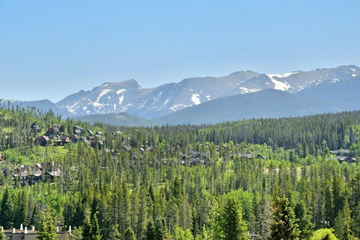 A sweeping view of snow-capped mountains and dense green trees, with scattered houses nestled in the valley. The landscape is peaceful and invites exploration, hinting at hiking, nature walks, and wildlife encounters. The changing colors of the sky suggest either sunrise or sunset, reinforcing the area’s year-round beauty.