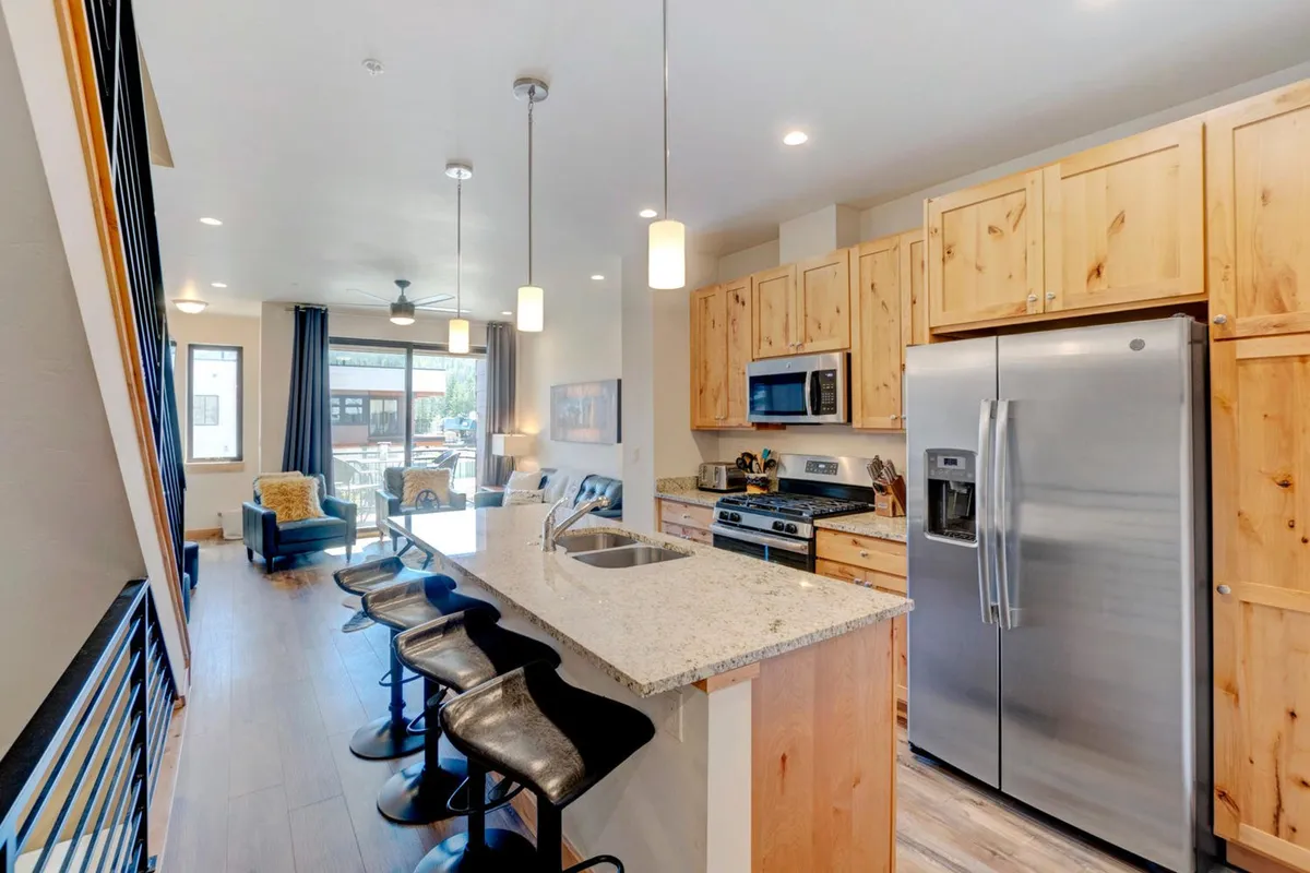 A warm, light-filled open kitchen featuring a large granite island with bar seating, stainless steel appliances, and expansive windows that fill the space with natural light. The island is the focal point, perfect for group cooking, casual meals, and socializing.
