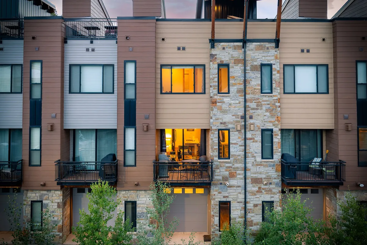 The exterior of the villa is shown with balconies and large windows aglow from warm interior lighting. The stone facade and surrounding environment evoke a welcoming, lively feeling, suggesting social gatherings and easy access to the downtown scene.