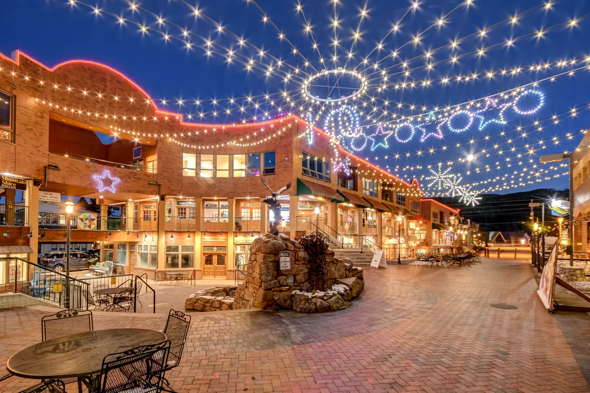 A cheerful outdoor space with decorative lights, a stone fountain, outdoor seating, and a brick walkway, evoking a sense of community and gathering.